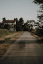 A rural road stretches into the distance, flanked by grassy fields and dense vegetation. A white house with a tiled roof is nestled among trees on the left. The scene is calm and serene, with scattered clouds in the sky.
