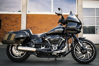 A sleek black motorcycle with chrome accents is parked in front of a brick wall, with a large exhaust and visible branding. The motorcycle has a modern design with sturdy tires and a streamlined body.