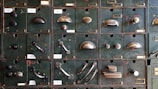 A collection of vintage metal drawer pulls and knobs mounted on an old, textured green panel. Each compartment features a different style and design of hardware, showcasing a variety of shapes and finishes.
