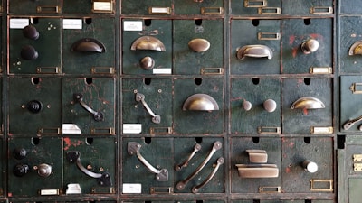 A collection of vintage metal drawer pulls and knobs mounted on an old, textured green panel. Each compartment features a different style and design of hardware, showcasing a variety of shapes and finishes.