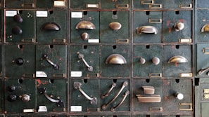 A collection of vintage metal drawer pulls and knobs mounted on an old, textured green panel. Each compartment features a different style and design of hardware, showcasing a variety of shapes and finishes.