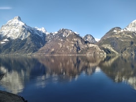 Snow-capped mountains reflect on a serene lake under a clear blue sky. The landscape is tranquil, with a mix of rocky peaks and wooded slopes. The stillness of the water enhances the peaceful atmosphere.
