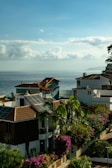 Coastal view with houses featuring red-tiled roofs and solar panels. Lush green palm trees and vibrant purple and pink bougainvillea bushes are prominent. The calm ocean stretches into the distance under a sky filled with fluffy, scattered clouds.