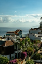 Coastal view with houses featuring red-tiled roofs and solar panels. Lush green palm trees and vibrant purple and pink bougainvillea bushes are prominent. The calm ocean stretches into the distance under a sky filled with fluffy, scattered clouds.
