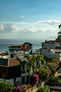 Coastal view with houses featuring red-tiled roofs and solar panels. Lush green palm trees and vibrant purple and pink bougainvillea bushes are prominent. The calm ocean stretches into the distance under a sky filled with fluffy, scattered clouds.