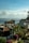 Coastal view with houses featuring red-tiled roofs and solar panels. Lush green palm trees and vibrant purple and pink bougainvillea bushes are prominent. The calm ocean stretches into the distance under a sky filled with fluffy, scattered clouds.