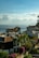 Coastal view with houses featuring red-tiled roofs and solar panels. Lush green palm trees and vibrant purple and pink bougainvillea bushes are prominent. The calm ocean stretches into the distance under a sky filled with fluffy, scattered clouds.