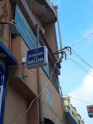 A street view featuring a building with various signs attached to it. One prominent sign reads 'Scissor The Salon' with an image of scissors above the text. The building appears weathered with visible power lines and pipes running along its exterior. A bright blue sky with scattered clouds is in the background, and part of the signage for another business is partially visible.