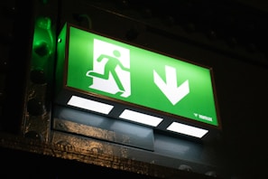 A green illuminated emergency exit sign displaying a pictogram of a person running towards an open door, alongside a downward-pointing arrow. The background around the sign is dark, highlighting the brightness of the sign.