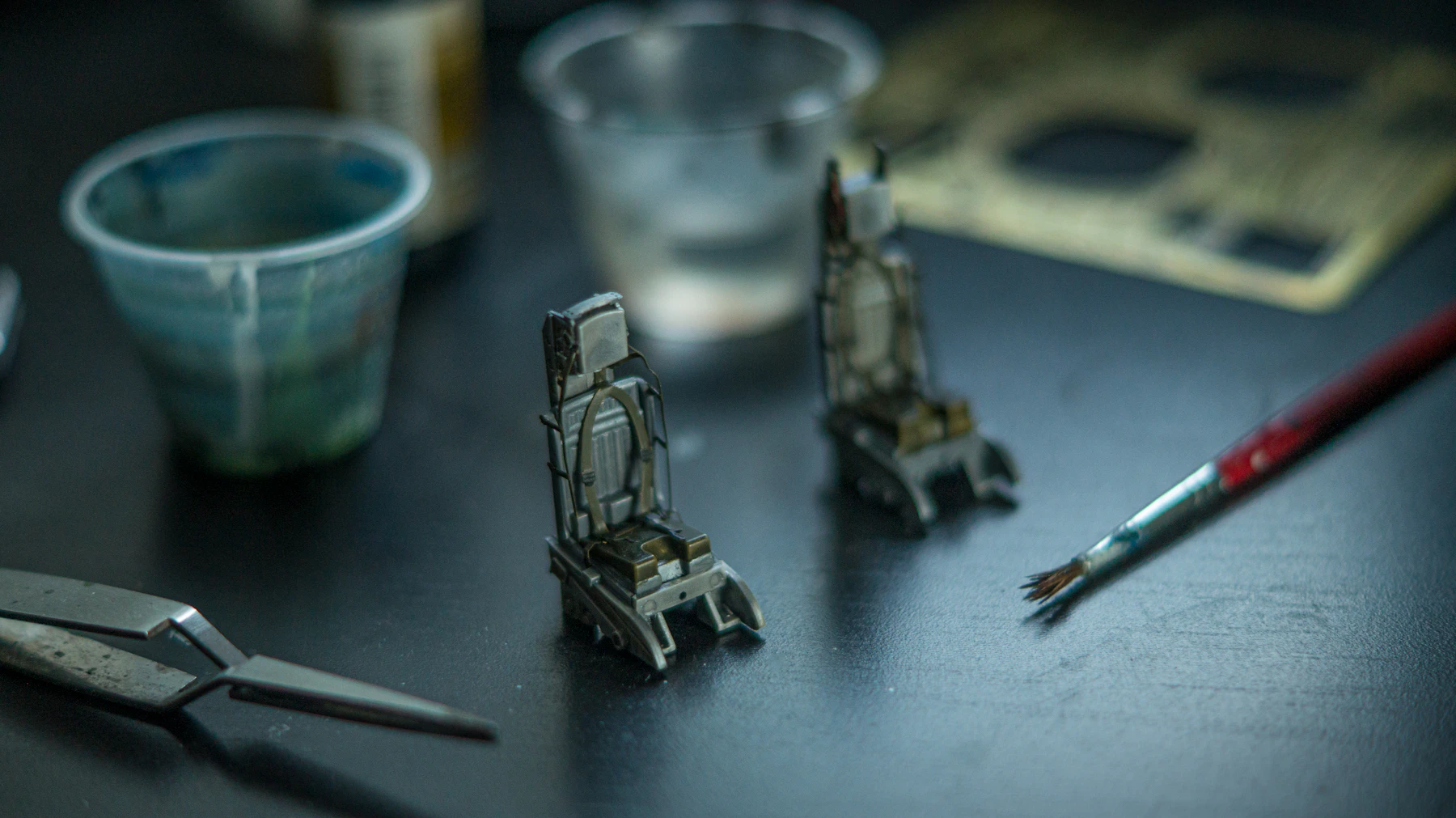 Two small gray model seats on a dark table beside plastic paint cups and a fine brush