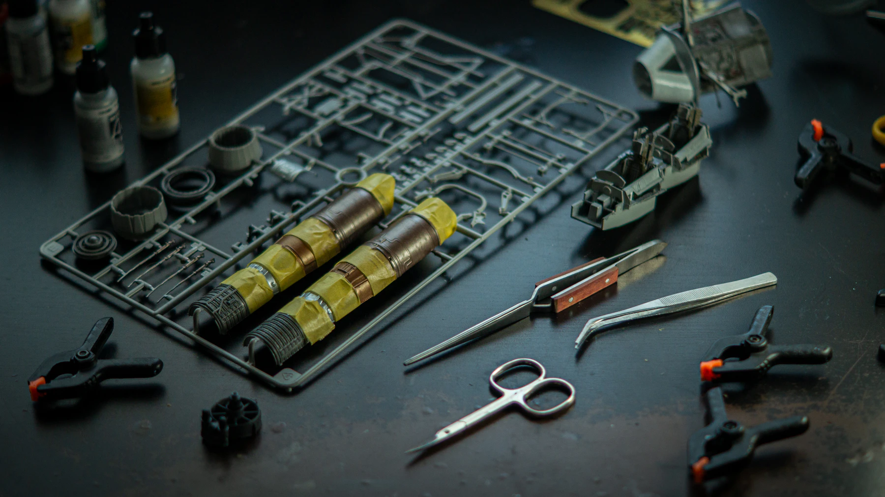 A gray plastic frame of kit parts on a black table beside tweezers, small clamps and paint bottles
