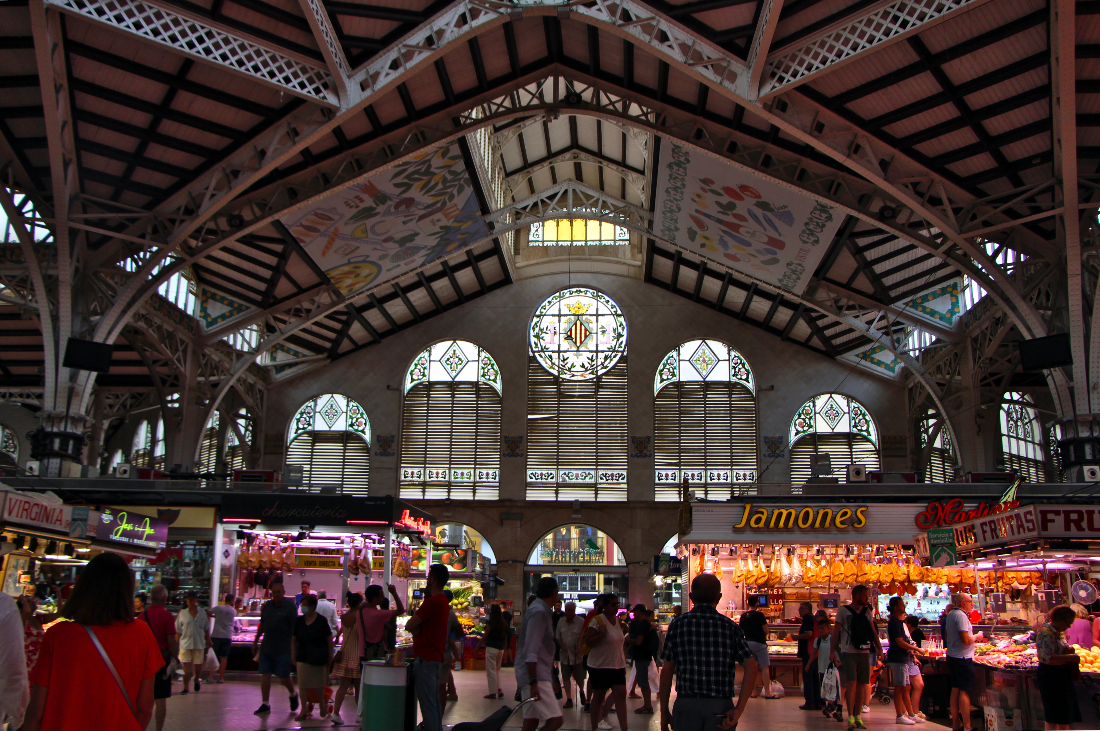 Shoppers beneath the iron-and-glass modernista vaults and stained glass of Valencia's Mercado Central, with a jamon stall in the foreground