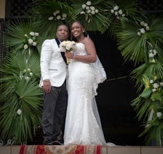 A couple dressed in formal wedding attire stands together. The woman is wearing a white lace wedding gown and holding a bouquet of flowers. The man is wearing a white suit jacket with dark trousers. They are positioned under a decorative arch made of green palm leaves and white flowers.