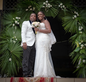 A couple dressed in formal wedding attire stands together. The woman is wearing a white lace wedding gown and holding a bouquet of flowers. The man is wearing a white suit jacket with dark trousers. They are positioned under a decorative arch made of green palm leaves and white flowers.