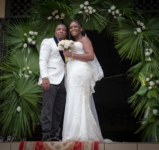 A couple dressed in formal wedding attire stands together. The woman is wearing a white lace wedding gown and holding a bouquet of flowers. The man is wearing a white suit jacket with dark trousers. They are positioned under a decorative arch made of green palm leaves and white flowers.