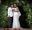 A couple dressed in formal wedding attire stands together. The woman is wearing a white lace wedding gown and holding a bouquet of flowers. The man is wearing a white suit jacket with dark trousers. They are positioned under a decorative arch made of green palm leaves and white flowers.