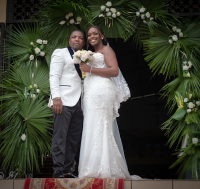 A couple dressed in formal wedding attire stands together. The woman is wearing a white lace wedding gown and holding a bouquet of flowers. The man is wearing a white suit jacket with dark trousers. They are positioned under a decorative arch made of green palm leaves and white flowers.