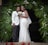 A couple dressed in formal wedding attire stands together. The woman is wearing a white lace wedding gown and holding a bouquet of flowers. The man is wearing a white suit jacket with dark trousers. They are positioned under a decorative arch made of green palm leaves and white flowers.