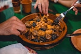 A ceramic dish filled with a variety of cooked foods such as apricots, nuts, and prunes, along with meat pieces, likely part of a traditional meal. Hands are reaching in, one holding a fork, suggesting communal dining.
