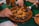 A ceramic dish filled with a variety of cooked foods such as apricots, nuts, and prunes, along with meat pieces, likely part of a traditional meal. Hands are reaching in, one holding a fork, suggesting communal dining.