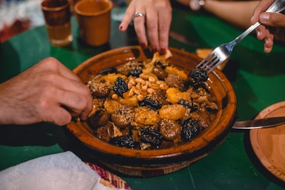 A ceramic dish filled with a variety of cooked foods such as apricots, nuts, and prunes, along with meat pieces, likely part of a traditional meal. Hands are reaching in, one holding a fork, suggesting communal dining.