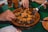 A ceramic dish filled with a variety of cooked foods such as apricots, nuts, and prunes, along with meat pieces, likely part of a traditional meal. Hands are reaching in, one holding a fork, suggesting communal dining.