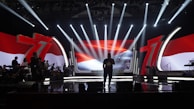 A stage with dramatic lighting and a large screen displaying a vibrant red and white background, featuring the number 71 prominently. Silhouetted figures stand across the stage, along with musical instruments, suggesting a performance setting.