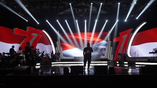 A stage with dramatic lighting and a large screen displaying a vibrant red and white background, featuring the number 71 prominently. Silhouetted figures stand across the stage, along with musical instruments, suggesting a performance setting.