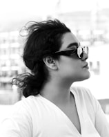 A black and white profile portrait of a person wearing sunglasses with a reflective surface. The individual has curly hair pulled back and is dressed in a white shirt. In the background, there's an out-of-focus urban setting with buildings, giving a sense of being outdoors.