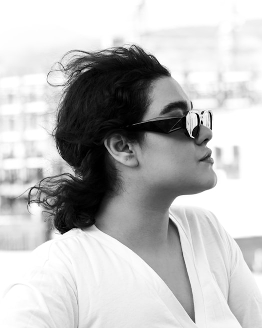 A black and white profile portrait of a person wearing sunglasses with a reflective surface. The individual has curly hair pulled back and is dressed in a white shirt. In the background, there's an out-of-focus urban setting with buildings, giving a sense of being outdoors.