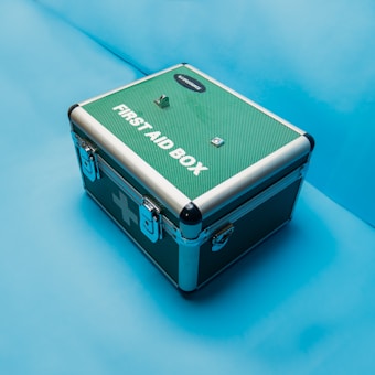 A green and silver first aid box with metal corners and locks is positioned on a solid light blue background. The box has prominent text on the top indicating its purpose.