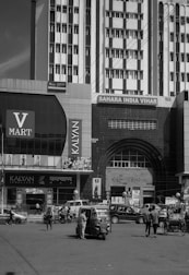 A busy urban street scene featuring a large commercial building with multiple signs, including V Mart and Kalyan Jewellers. Several vehicles, including an auto rickshaw and pedestrians, are visible in front of the building. The architecture includes tall structures with numerous windows and an arched entrance labeled 'RAJ TOWER'.