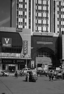 A busy urban street scene featuring a large commercial building with multiple signs, including V Mart and Kalyan Jewellers. Several vehicles, including an auto rickshaw and pedestrians, are visible in front of the building. The architecture includes tall structures with numerous windows and an arched entrance labeled 'RAJ TOWER'.