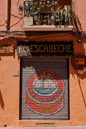 A street scene with a closed storefront featuring a metallic roll-up door decorated with a circular logo that includes an illustration of a ship on the sea. Above the door, there is a small balcony with several potted plants and a few warning signs about noise. The building exterior is painted in a warm terracotta color.