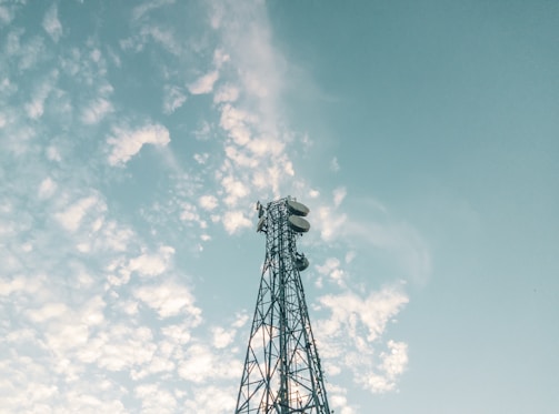 A tall metal telecommunications tower with several satellite dishes and antennas stands against a sky filled with scattered white clouds. The structure extends upwards towards the clear, light blue backdrop, conveying a sense of height and open space.