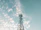 A tall metal telecommunications tower with several satellite dishes and antennas stands against a sky filled with scattered white clouds. The structure extends upwards towards the clear, light blue backdrop, conveying a sense of height and open space.