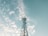 A tall metal telecommunications tower with several satellite dishes and antennas stands against a sky filled with scattered white clouds. The structure extends upwards towards the clear, light blue backdrop, conveying a sense of height and open space.