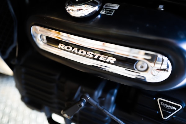 Close-up of a motorcycle engine part featuring a metallic panel with the word 'ROADSTER' prominently displayed. The engine has a sleek black finish with chrome accents. The Triumph logo is visible on another part of the engine.