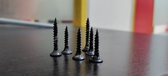 A close-up view of several black screws standing upright on a dark surface, with a blurred background featuring vertical bands of red and yellow.
