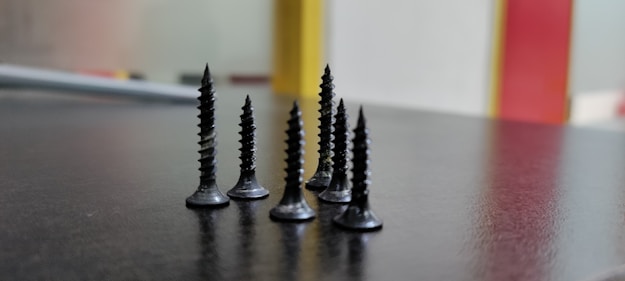A close-up view of several black screws standing upright on a dark surface, with a blurred background featuring vertical bands of red and yellow.