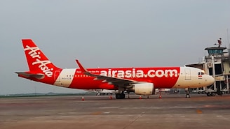 A red and white AirAsia airplane is parked on the tarmac near an airport terminal. The aircraft displays the airline's logo prominently on its fuselage. The scene appears to be set in a cloudy environment, adding a sense of overcast lighting to the setting.