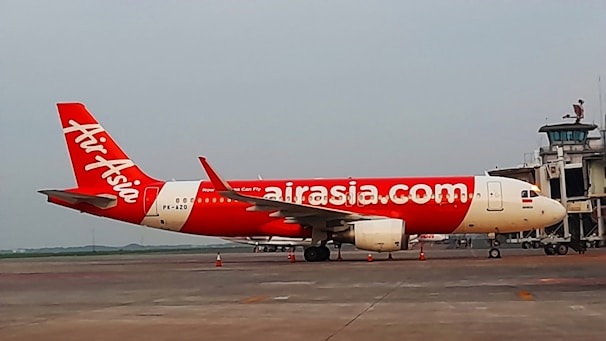 A red and white AirAsia airplane is parked on the tarmac near an airport terminal. The aircraft displays the airline's logo prominently on its fuselage. The scene appears to be set in a cloudy environment, adding a sense of overcast lighting to the setting.