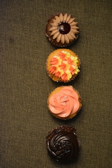 Four cupcakes are neatly arranged in a vertical line on a textured green surface. Each cupcake has distinct frosting designs and colors. The top cupcake has chocolate frosting with a central glossy decoration, resembling a flower or sun. The second cupcake features orange and yellow star-like icing patterns. The third cupcake displays a swirl of pink frosting adorned with small colorful sprinkles. The bottom cupcake has a richly dark chocolate frosting with a glossy finish.