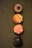 Four cupcakes are neatly arranged in a vertical line on a textured green surface. Each cupcake has distinct frosting designs and colors. The top cupcake has chocolate frosting with a central glossy decoration, resembling a flower or sun. The second cupcake features orange and yellow star-like icing patterns. The third cupcake displays a swirl of pink frosting adorned with small colorful sprinkles. The bottom cupcake has a richly dark chocolate frosting with a glossy finish.