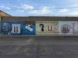 A long mural painted on a brick wall features various symbols and logos representing different sports clubs. The mural includes vivid portraits and emblems, with each section dedicated to a specific team. There are distinctive club colors and badges, complemented by text and artistic elements.