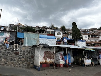 A hillside view of a village with various small houses and market stalls. The structures are made of mixed materials, including tarps and metal sheets. In the foreground, two people are sitting on a stone wall. The sky is overcast with gray clouds, and there are trees interspersed among the buildings.