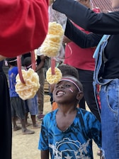 A group of children is participating in a game where snacks are tied to strings and hung from above. One child is looking up at a piece of food hanging in front of him, wearing a blue shirt with a pattern. Another child in the background is waiting for their turn. Some adults are holding the strings. Flags and festive decorations are visible, indicating a celebration or event.