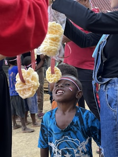 A group of children is participating in a game where snacks are tied to strings and hung from above. One child is looking up at a piece of food hanging in front of him, wearing a blue shirt with a pattern. Another child in the background is waiting for their turn. Some adults are holding the strings. Flags and festive decorations are visible, indicating a celebration or event.