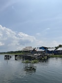 A rural scene featuring a row of wooden houses built over a body of water, likely a lake or river. The homes have rustic, weathered structures with corrugated metal roofs. Vegetation is visible both on the water and in the background, including trees under a bright, partly cloudy sky.