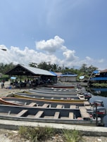 A row of small wooden boats with outboard motors is aligned on a concrete ramp by the water's edge. The area is shaded by a large roofed structure where a group of people is gathered. A clear blue sky with scattered fluffy clouds forms the backdrop, and lush green trees are visible in the background.